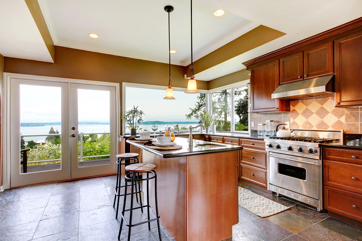 A modern kitchen features dark wood cabinetry, a large island with seating, and stainless steel appliances, with scenic views of a lake and greenery visible through large windows and doors.