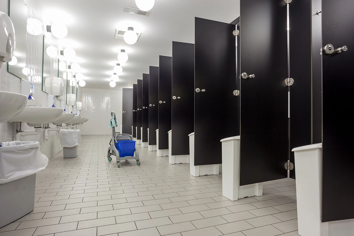 A clean public restroom featuring several black stall doors, sinks with blue soap dispensers, and a cleaning cart in the center. Bright overhead lights illuminate the tiled floor.