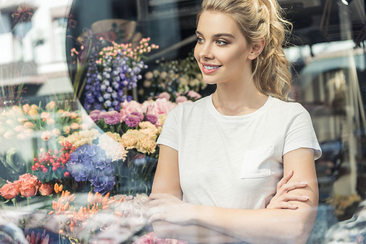 A smiling woman in a white t-shirt stands with her arms crossed in front of a flower shop, surrounded by an array of colorful blooms.