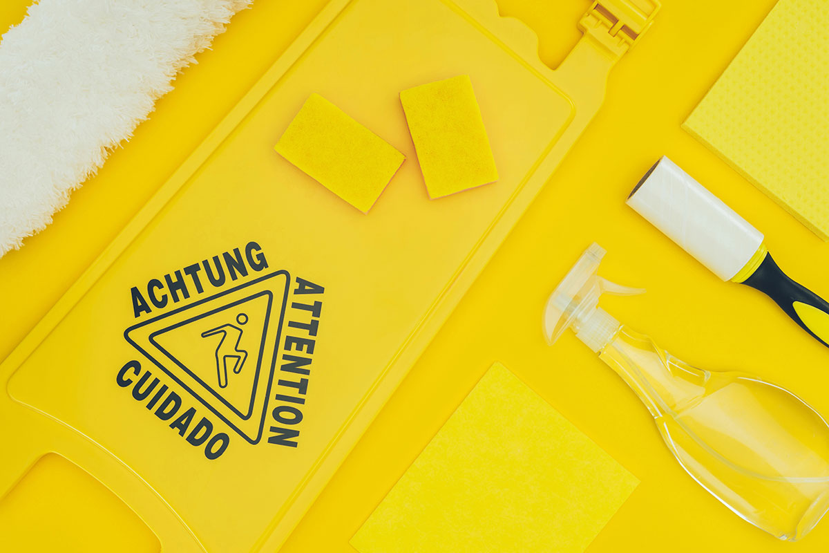 A flat lay of cleaning supplies on a bright yellow background, including two yellow sponges, a caution sign with multilingual warnings, a lint brush, a spray bottle, and two yellow cleaning cloths.