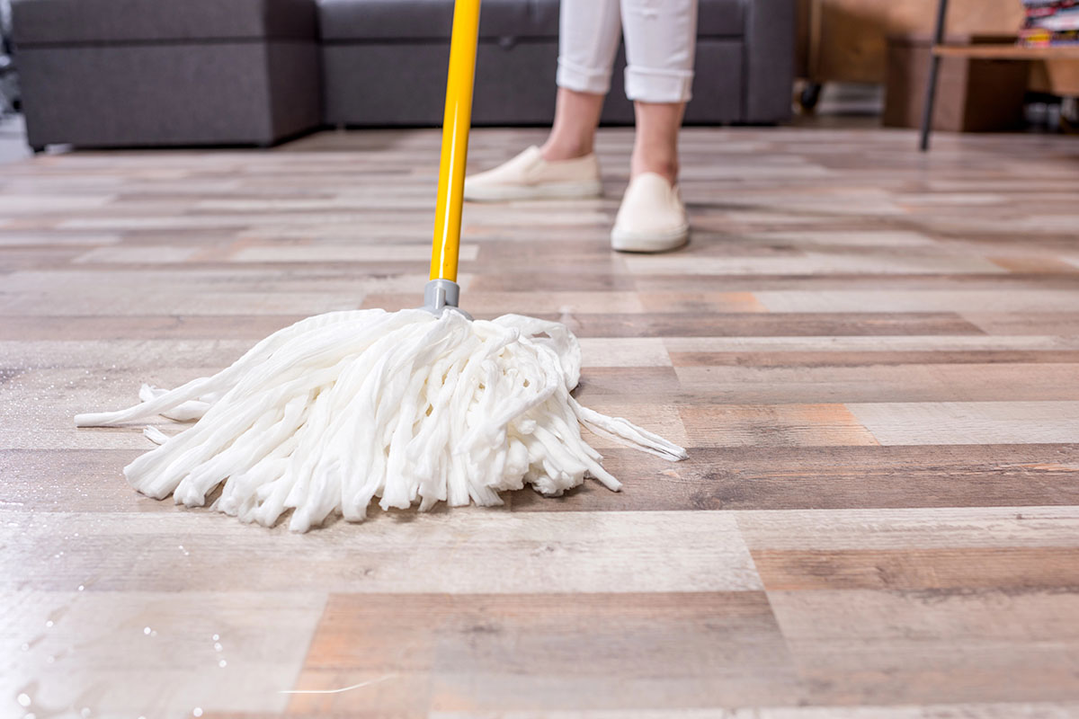 A person mopping a wooden floor with a white mop and a yellow handle, wearing white pants and slip-on shoes.